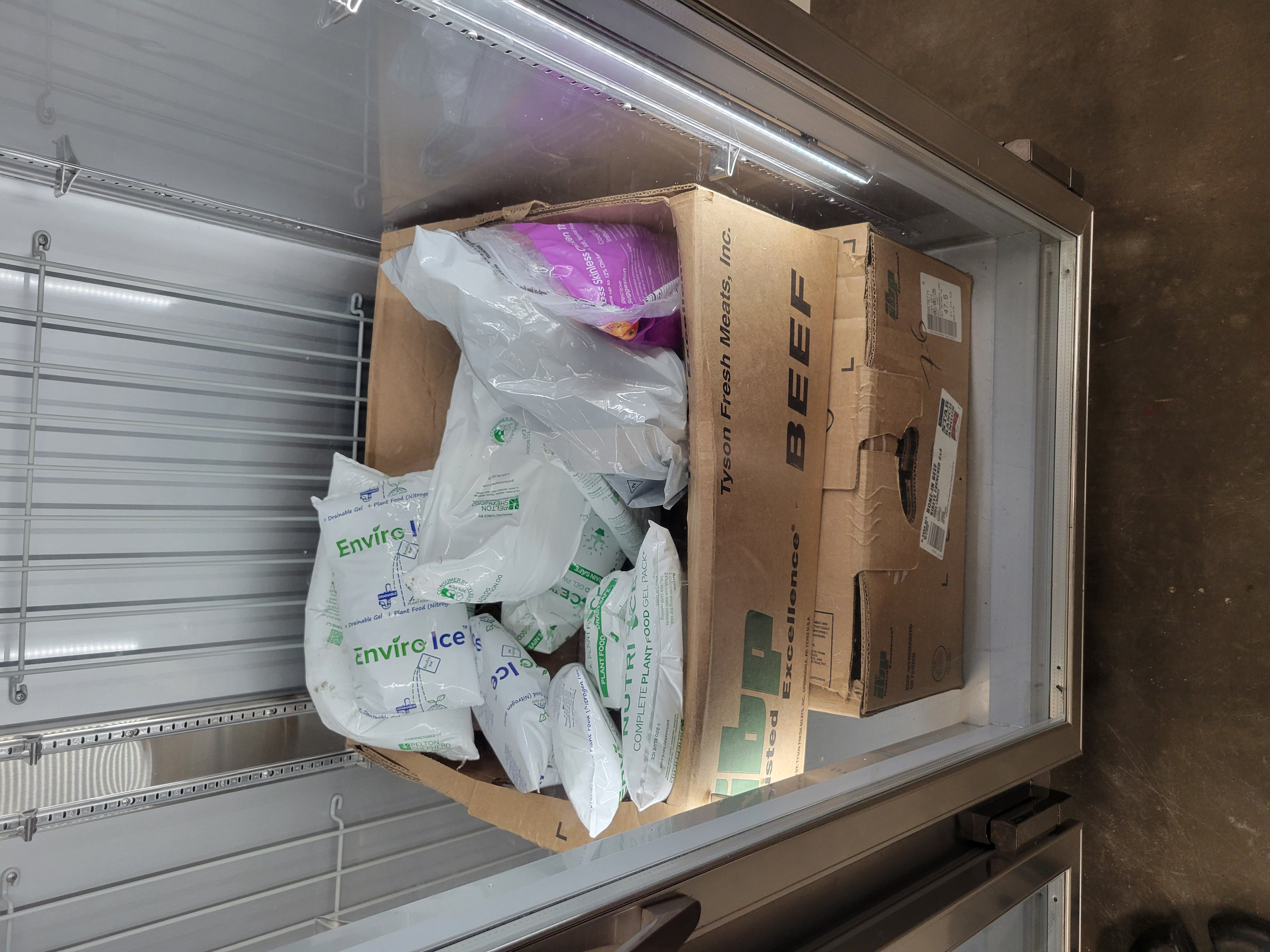 Food gel packs with a bag of skinless chicken tighs in a cardboard box on top of another box in a fridge